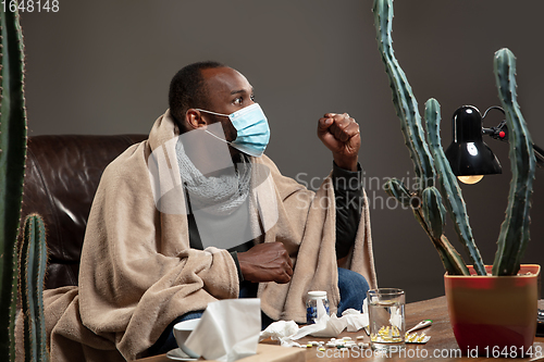 Image of Young man wrapped in a plaid looks sick, ill, sneezing and coughing sitting at home indoors in face mask.