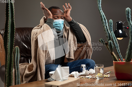 Image of Young man wrapped in a plaid looks sick, ill, sneezing and coughing sitting at home indoors in face mask.