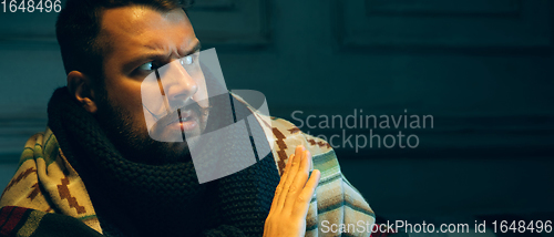 Image of Man wrapped in a plaid looks sick, ill, sneezing and coughing sitting at home indoors