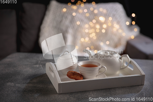 Image of Cozy winter. White wooden tray with tea set on grey table with led garland lights. The concept of home atmosphere and comfort.