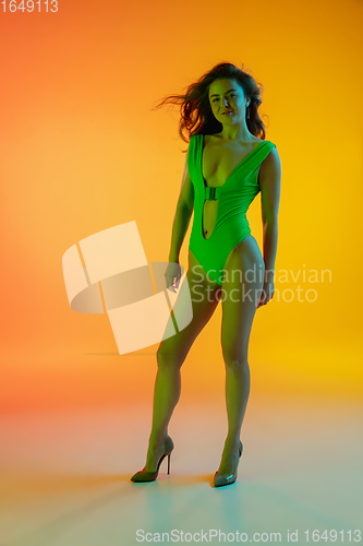 Image of Beautiful seductive girl in fashionable green swimsuit on bright gradient yellow-orange background in neon light