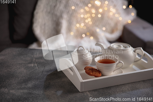 Image of Cozy winter. White wooden tray with tea set on grey table with led garland lights. The concept of home atmosphere and comfort.