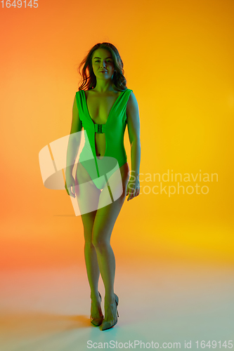 Image of Beautiful seductive girl in fashionable green swimsuit on bright gradient yellow-orange background in neon light