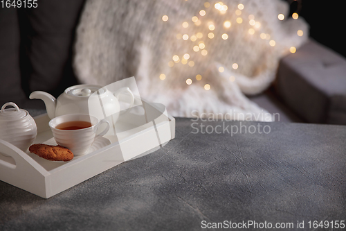 Image of Cozy winter. White wooden tray with tea set on grey table with led garland lights. The concept of home atmosphere and comfort.