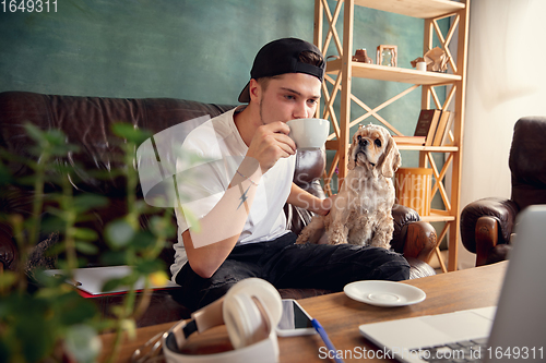 Image of Young handsome man sitting on sofa at home with his cute dog. Cozy office workplace, remote work, online, e-learning concept.