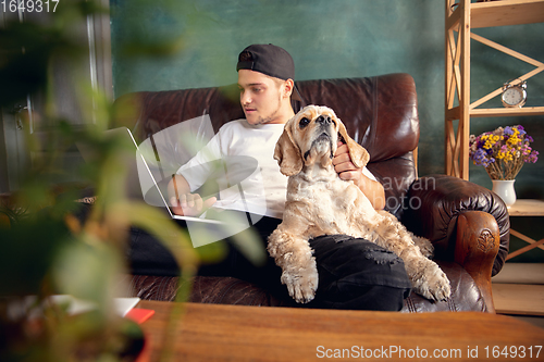 Image of Young handsome man sitting and working at home with his cute dog. Cozy office workplace, remote work, online learning concept.