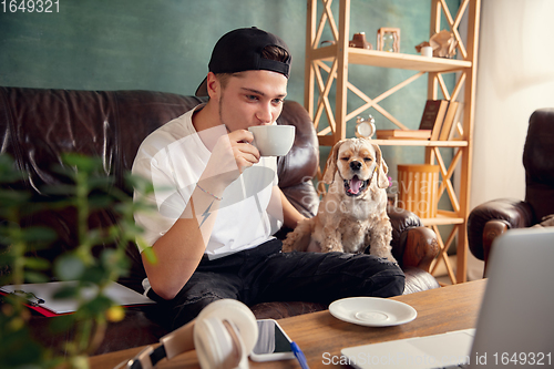 Image of Young handsome man sitting on sofa at home with his cute dog. Cozy office workplace, remote work, online, e-learning concept.