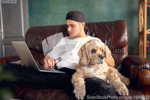 Image of Young handsome man sitting and working at home with his cute dog. Cozy office workplace, remote work, online learning concept.