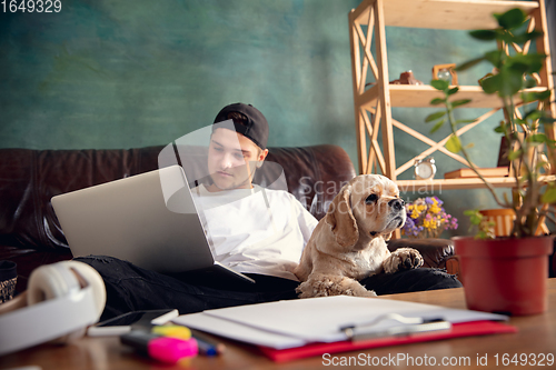 Image of Young handsome man sitting and working at home with his cute dog. Cozy office workplace, remote work, online learning concept.