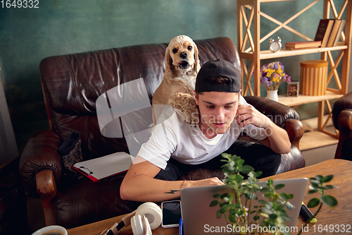 Image of Young man sitting on sofa at home with his cute dog. Cozy office workplace, remote work, online, e-learning concept.