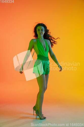 Image of Beautiful seductive girl in fashionable, summer outfit, green jumpsuit on bright gradient yellow-orange background in neon light