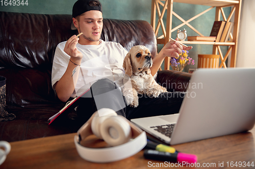 Image of Young man sitting on sofa at home with cute dog and chatting with his friends online.