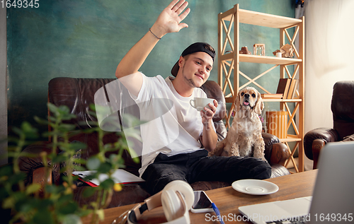 Image of Young man sitting on sofa at home with cute dog and chatting with his friends online.