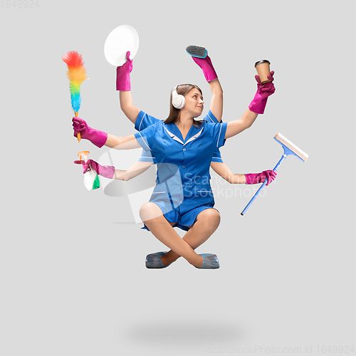 Image of Young woman housemaid in headphones and blue uniform multitask like shiva isolated on gray background.
