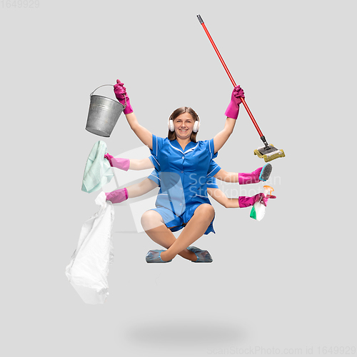 Image of Young woman housemaid in headphones and blue uniform multitask like shiva isolated on gray background.