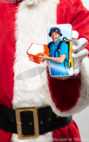 Image of Close up hands of Santa Claus holding device with happy deliveryman on the screen