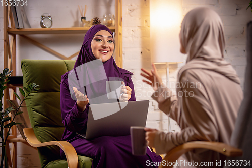 Image of Happy two muslim women at home during lesson, studying with devices, online or home education