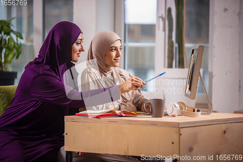 Image of Happy two muslim women at home during lesson, studying near computer, online education