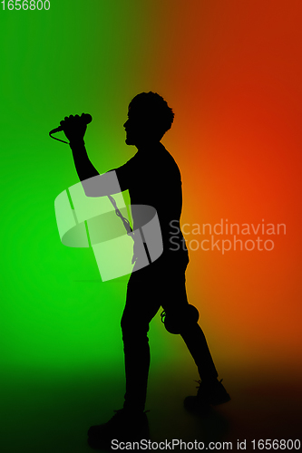 Image of Silhouette of young caucasian male singer isolated on green-orange gradient studio background in neon light