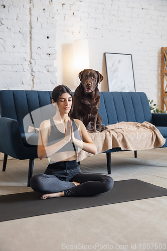 Image of Young woman working out at home during lockdown, yoga exercises with the dog