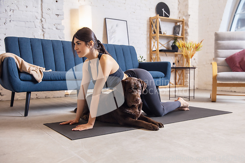 Image of Young woman working out at home during lockdown, yoga exercises with the dog