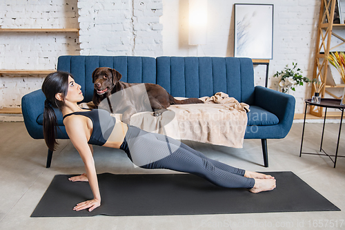 Image of Young woman working out at home during lockdown, yoga exercises with the dog