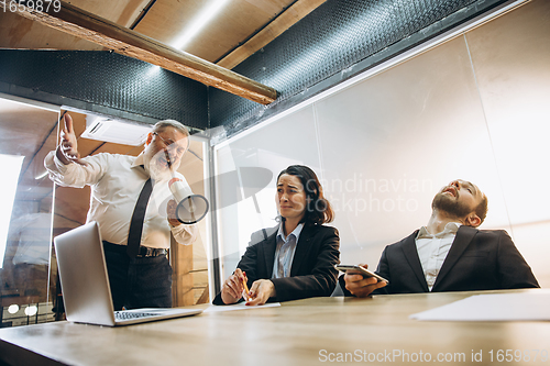 Image of Angry boss with megaphone screaming at employees in office, scared and annoyed colleagues listening at the table