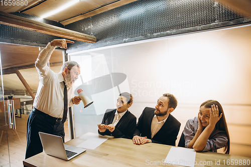 Image of Angry boss with megaphone screaming at employees in office, scared and annoyed colleagues listening at the table