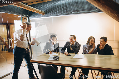 Image of Angry boss with megaphone screaming at employees in office, scared and annoyed colleagues listening at the table