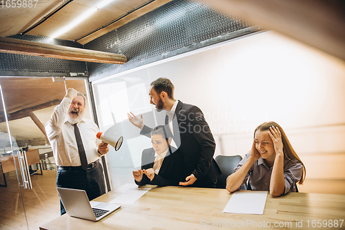 Image of Angry boss with megaphone screaming at employees in office, scared and annoyed colleagues listening at the table