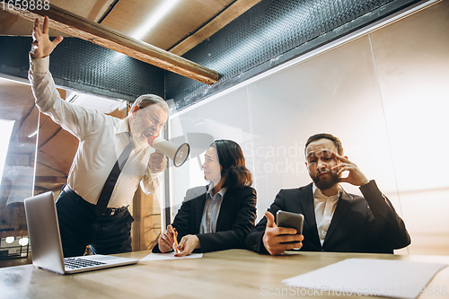 Image of Angry boss with megaphone screaming at employees in office, scared and annoyed colleagues listening at the table