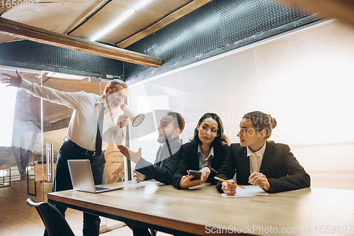 Image of Angry boss with megaphone screaming at employees in office, scared and annoyed colleagues listening at the table