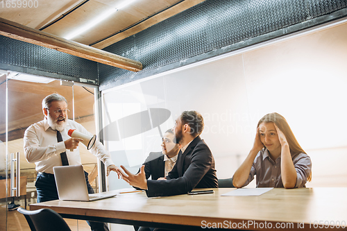 Image of Angry boss with megaphone screaming at employees in office, scared and annoyed colleagues listening at the table