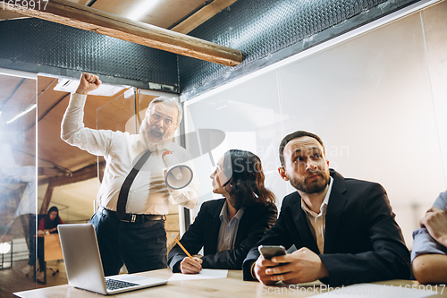 Image of Angry boss with megaphone screaming at employees in office, scared and annoyed colleagues listening at the table