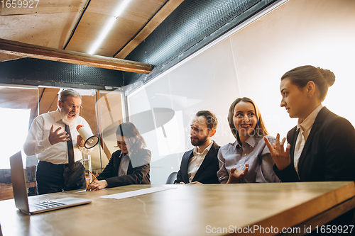 Image of Angry boss with megaphone screaming at employees in office, scared and annoyed colleagues listening at the table