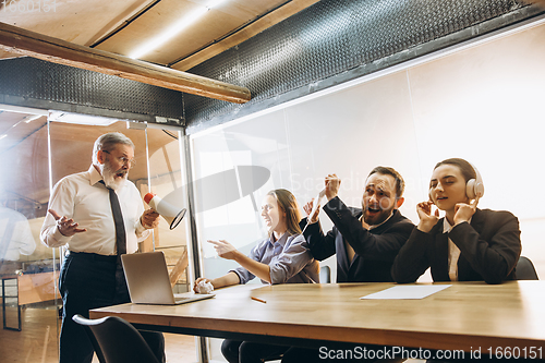 Image of Angry boss with megaphone screaming at employees in office, scared and annoyed colleagues listening at the table