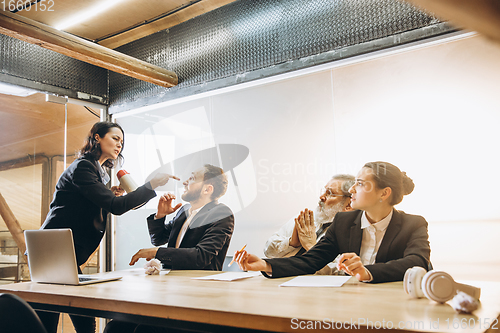 Image of Angry boss with megaphone screaming at employees in office, scared and annoyed colleagues listening at the table