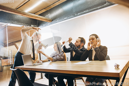 Image of Angry boss with megaphone screaming at employees in office, scared and annoyed colleagues listening at the table