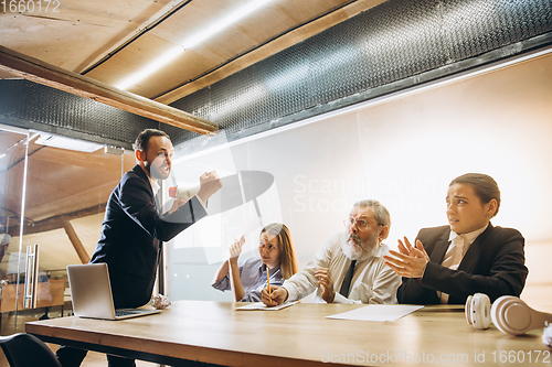 Image of Angry boss with megaphone screaming at employees in office, scared and annoyed colleagues listening at the table