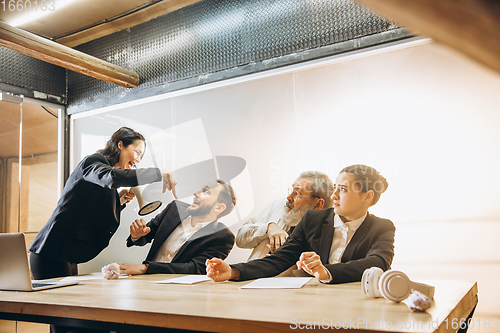 Image of Angry boss with megaphone screaming at employees in office, scared and annoyed colleagues listening at the table