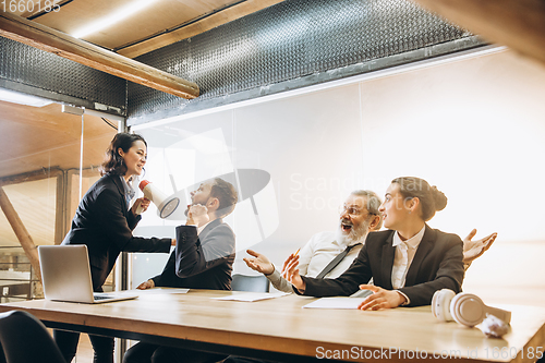 Image of Angry boss with megaphone screaming at employees in office, scared and annoyed colleagues listening at the table