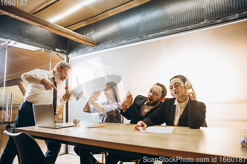 Image of Angry boss with megaphone screaming at employees in office, scared and annoyed colleagues listening at the table