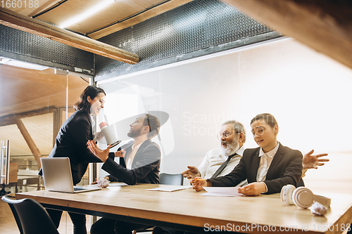 Image of Angry boss with megaphone screaming at employees in office, scared and annoyed colleagues listening at the table