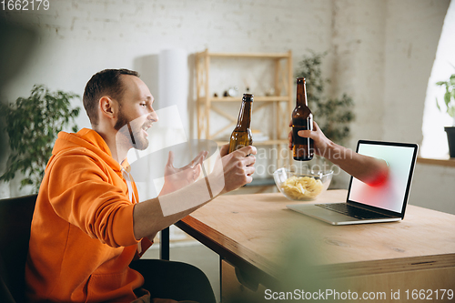 Image of Young man drinking beer during meeting friends on virtual video call. Distance online meeting, chat together on laptop at home.