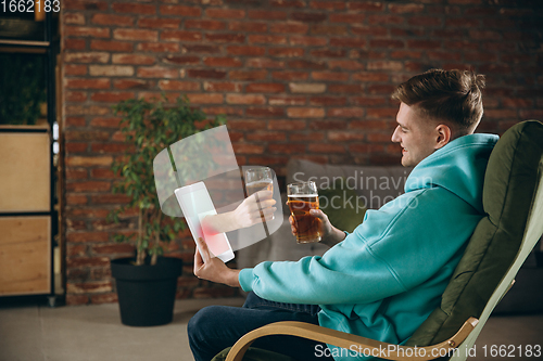 Image of Young man drinking beer during meeting friends on virtual video call. Distance online meeting, chat together on tablet at home.