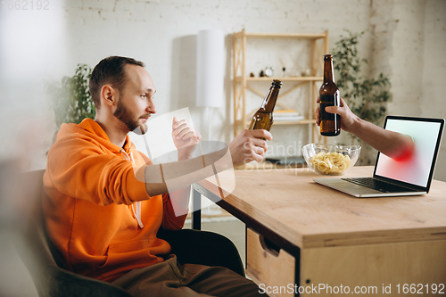 Image of Young man drinking beer during meeting friends on virtual video call. Distance online meeting, chat together on laptop at home.