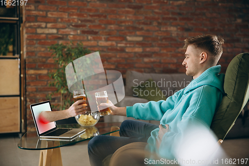 Image of Young man drinking beer during meeting friends on virtual video call. Distance online meeting, chat together on laptop at home.