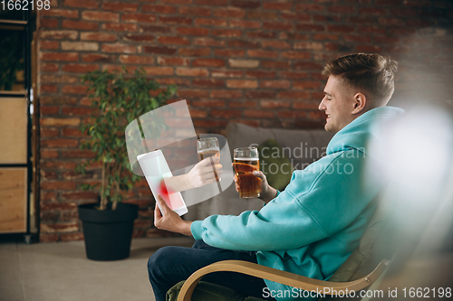 Image of Young man drinking beer during meeting friends on virtual video call. Distance online meeting, chat together on tablet at home.