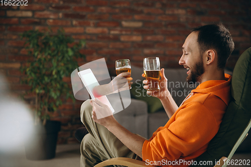 Image of Young man drinking beer during meeting friends on virtual video call. Distance online meeting, chat together on tablet at home.