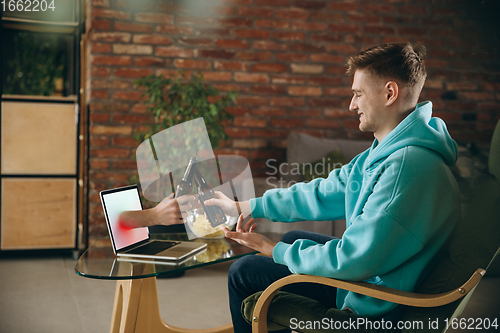 Image of Young man drinking beer during meeting friends on virtual video call. Distance online meeting, chat together on laptop at home.